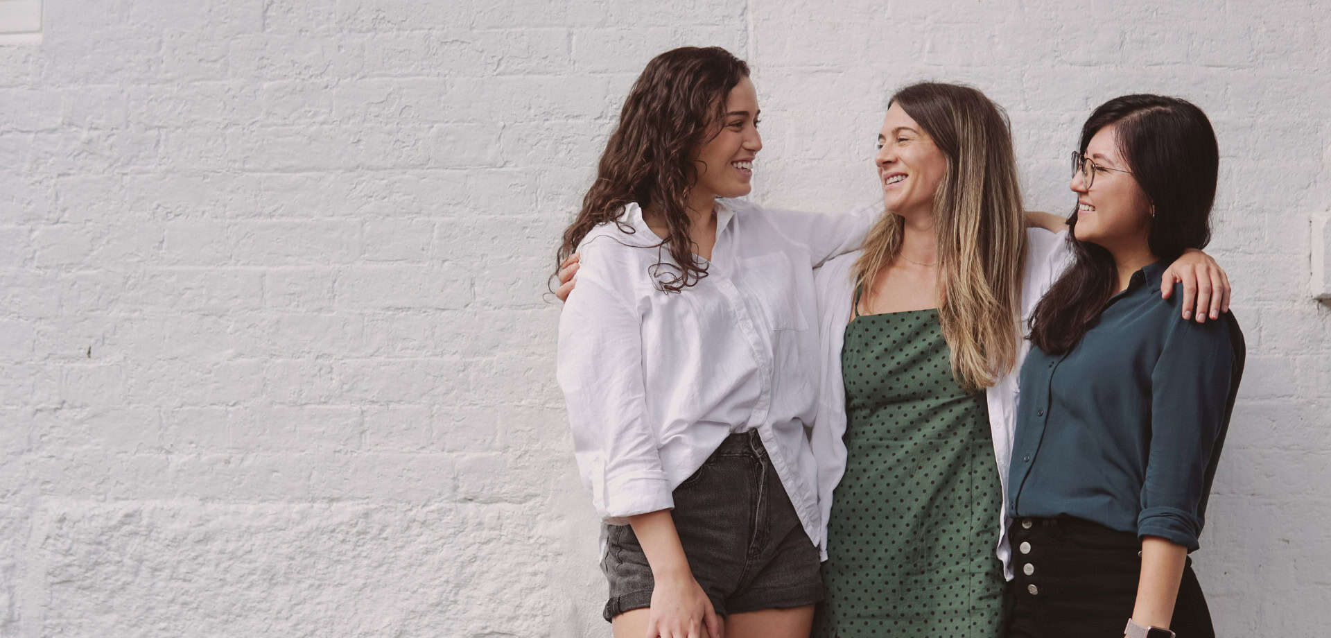 A group of three women laughing and having a conversation against a white brick wall background.