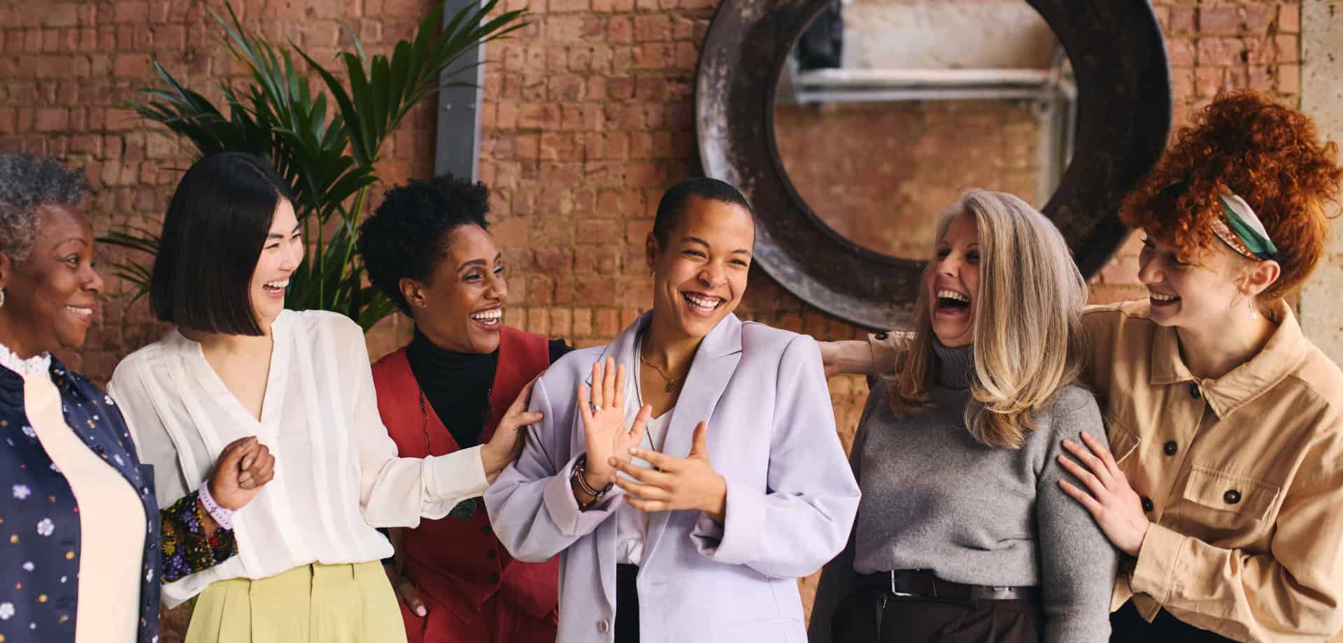 A group of six women laughing and joking with each other with a stone wall background.